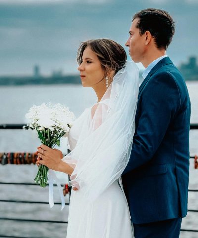 A bride and groom pose elegantly by the waterfront on a cloudy day.